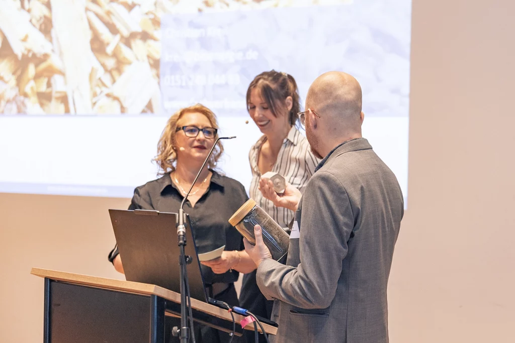 Yvonne Bosch (Bundesgütegemeinschaft Holzasche e.V.) und Anna-Marlen Vöcking (Technologica GmbH) übergeben ein Geschenk an einen Referenten auf dem Holzaschekongress 2026.